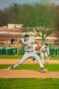 High school baseball player wearing a Tigers uniform throws a pitch on the mound during a sunny day game.