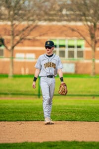 Baseball player in gray uniform walking on the field, preparing for the game. Green grass and trees in the background.