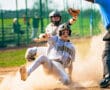 Youth baseball player slides into home plate, kicking up dust as the umpire watches the action unfold.