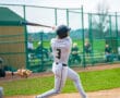 Young baseball player in uniform swings bat at outdoor field, aiming for the ball during a game. Spectators in background.