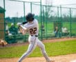 Baseball player in white uniform swings bat at the ball during a game, with spectators in the background on a sunny day.