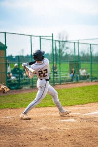 Baseball player in white uniform swings bat at the ball during a game, with spectators in the background on a sunny day.