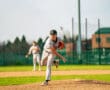 A young pitcher throws a baseball on a sunny day, with a teammate in the background on a grassy baseball field.