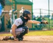 A youth baseball catcher signals for a pitch, wearing protective gear on a sunny field. Team members are in the background.