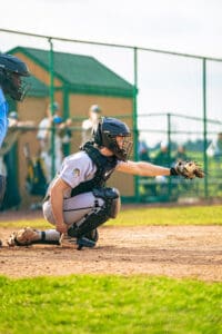 A youth baseball catcher signals for a pitch, wearing protective gear on a sunny field. Team members are in the background.