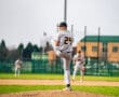 High school baseball pitcher in action, delivering a pitch on the mound during a game. Teammate visible in the background.