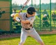 Young baseball player in a gray uniform stands ready to bat during a game, with a fence and spectators in the background.