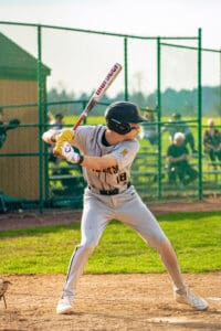 Young baseball player in a gray uniform stands ready to bat during a game, with a fence and spectators in the background.