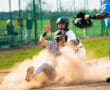 A youth baseball player slides into home plate as the catcher prepares to tag him, with dust flying around the action.