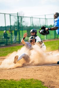 A youth baseball player slides into home plate as the catcher prepares to tag him, with dust flying around the action.