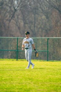 A baseball player in a gray uniform walks on a green field, wearing sunglasses and a glove, ready for the game.