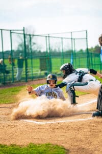 A young baseball player slides into home plate as the catcher prepares for the play, dust swirling around them.