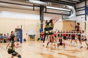 Boys volleyball match in action, players jumping at the net while spectators watch enthusiastically.