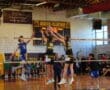 Two volleyball players jump to block a spike during a match at Huston-Tillotson University, with spectators watching.