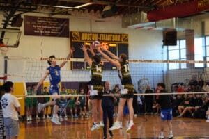 Two volleyball players jump to block a spike during a match at Huston-Tillotson University, with spectators watching.