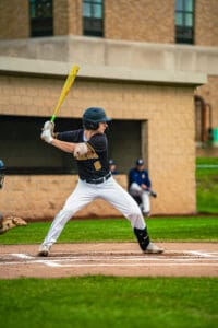 A young baseball player at bat, poised to swing, on a green field during a game. Teammates are visible in the background.