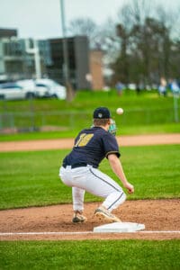 A baseball player crouches at first base, ready to catch a ball during a game on a grassy field.