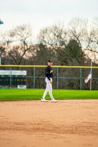 Baseball player in uniform walking on the field during practice, with trees and an American flag in the background.