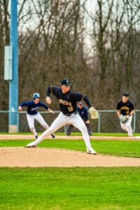 High school baseball pitcher winding up to throw a ball, with players running in the background on a grassy field.
