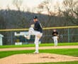 High school baseball pitcher in a black jersey stands on the mound, ready to deliver a pitch during a game.