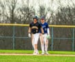 Two baseball players chat on the field, wearing team jerseys and helmets, during a game on a sunny day.