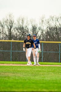 Two baseball players chat on the field, wearing team jerseys and helmets, during a game on a sunny day.