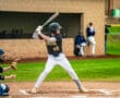 A young baseball player in a black jersey prepares to swing a bat during a game, while a catcher awaits the pitch.
