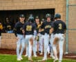 High school baseball players celebrating a play, wearing black jerseys and chatting on the field near the dugout.