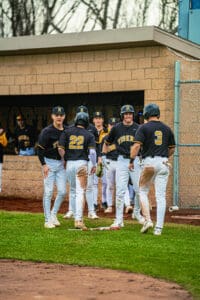 High school baseball players celebrating a play, wearing black jerseys and chatting on the field near the dugout.