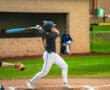 Young baseball player swings a bat during a game, while teammates observe from the dugout in the background.