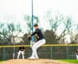 A young baseball pitcher winds up on the mound during a game; teammates position themselves in the background.