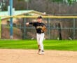 Young baseball player in a black team uniform throws the ball from the infield during practice on a sunny day.