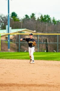 Young baseball player in a black team uniform throws the ball from the infield during practice on a sunny day.
