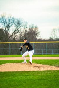 A young baseball pitcher in mid-throw on the mound, wearing a black jersey and white pants in a grassy field.