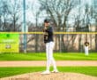 Young male baseball pitcher stands on the mound, preparing to pitch in a game, with teammates in the background.