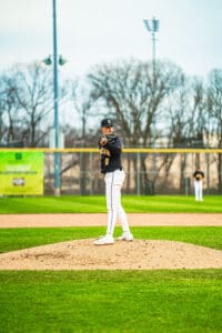 Young male baseball pitcher stands on the mound, preparing to pitch in a game, with teammates in the background.