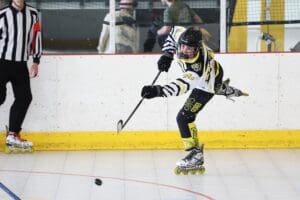 Young athlete skating and shooting a puck in a roller hockey game, with a referee in the background for officiating.