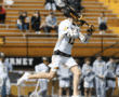 Lacrosse player in action, shooting on goal during a sunny game day with fans in the background on yellow bleachers.