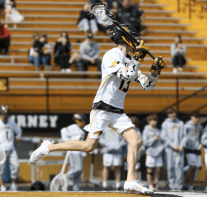 Lacrosse player in action, shooting on goal during a sunny game day with fans in the background on yellow bleachers.