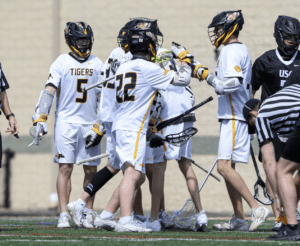 Lacrosse players in white uniforms celebrate together on the field during a game, showcasing teamwork and excitement.