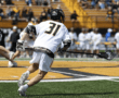 Player in white lacrosse uniform runs on a grassy field, stick in hand, during an outdoor game with spectators in the background.