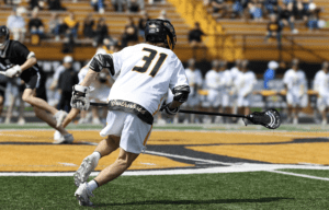 Player in white lacrosse uniform runs on a grassy field, stick in hand, during an outdoor game with spectators in the background.