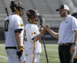 Lacrosse players in team jerseys engage in a motivational huddle with their coach on the field.