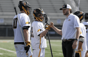 Lacrosse players in team jerseys engage in a motivational huddle with their coach on the field.