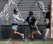 Lacrosse players competing as one attempts a shot while the goalie prepares to block it during an outdoor game.