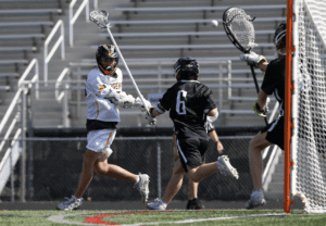 Lacrosse players competing as one attempts a shot while the goalie prepares to block it during an outdoor game.