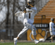 A lacrosse player in action, wearing a white jersey and gray shorts, throwing a ball on a sunny day.