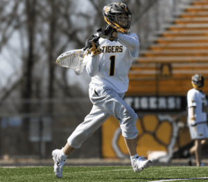 A lacrosse player in action, wearing a white jersey and gray shorts, throwing a ball on a sunny day.