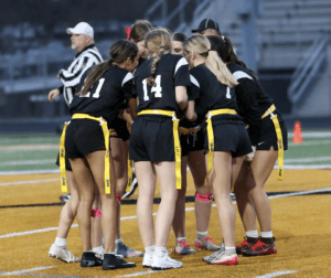 A group of female players in black jerseys huddle on a football field, preparing for their flag football game.
