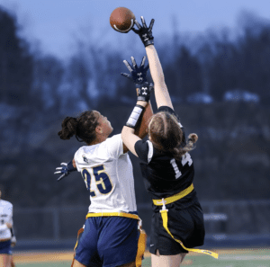 Two female athletes compete for a football in a dynamic moment during a flag football game under dim lights.